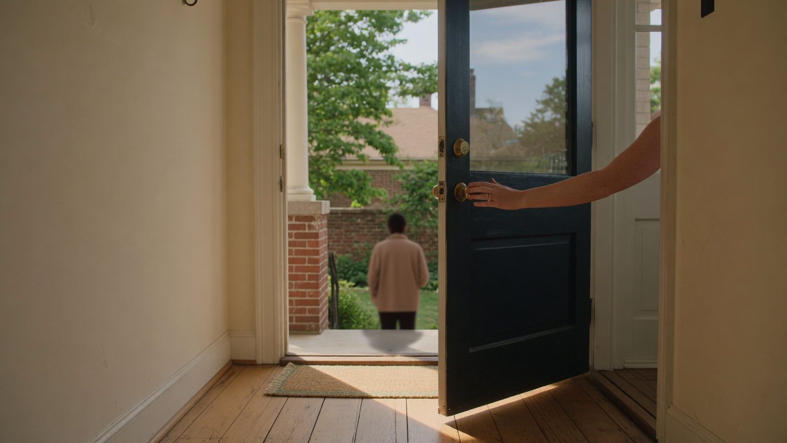 Homeowner's hand on a front door opened slightly onto a porch in daylight.